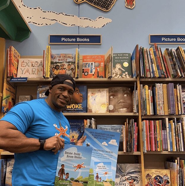 A man wearing a blue tshirt and holding books with a lot of books in the background